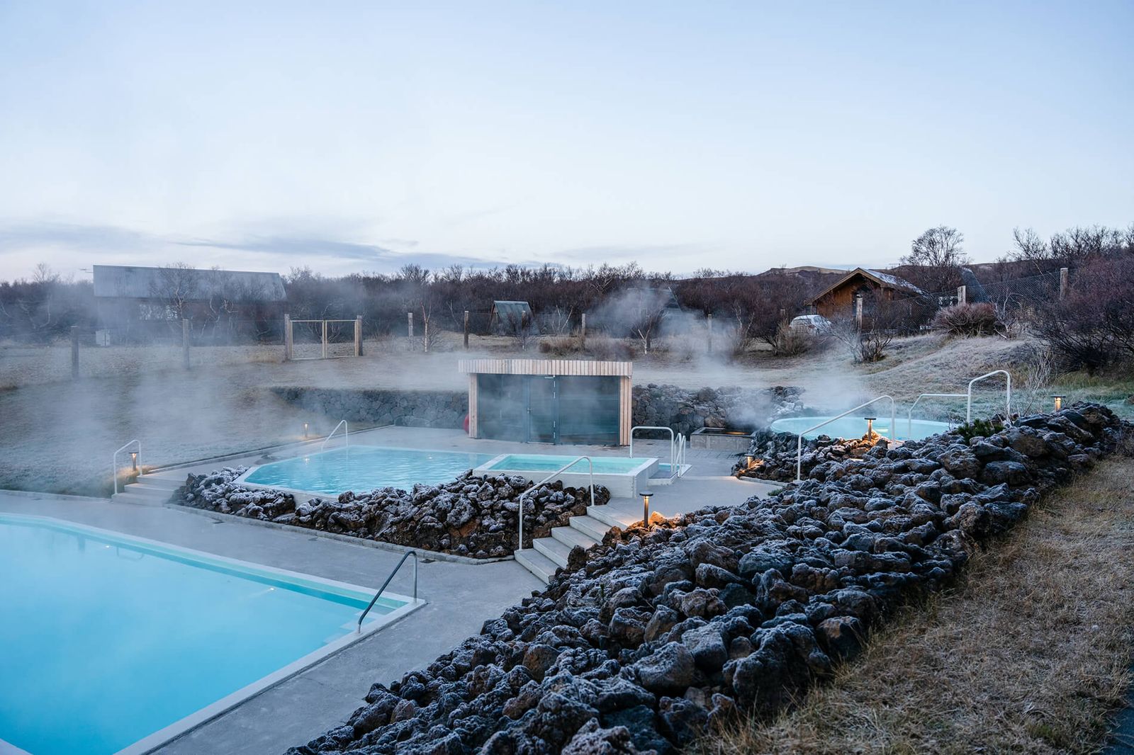 Steam rising from outdoor geothermal pools surrounded by dark lava rock and frost at Húsafell.