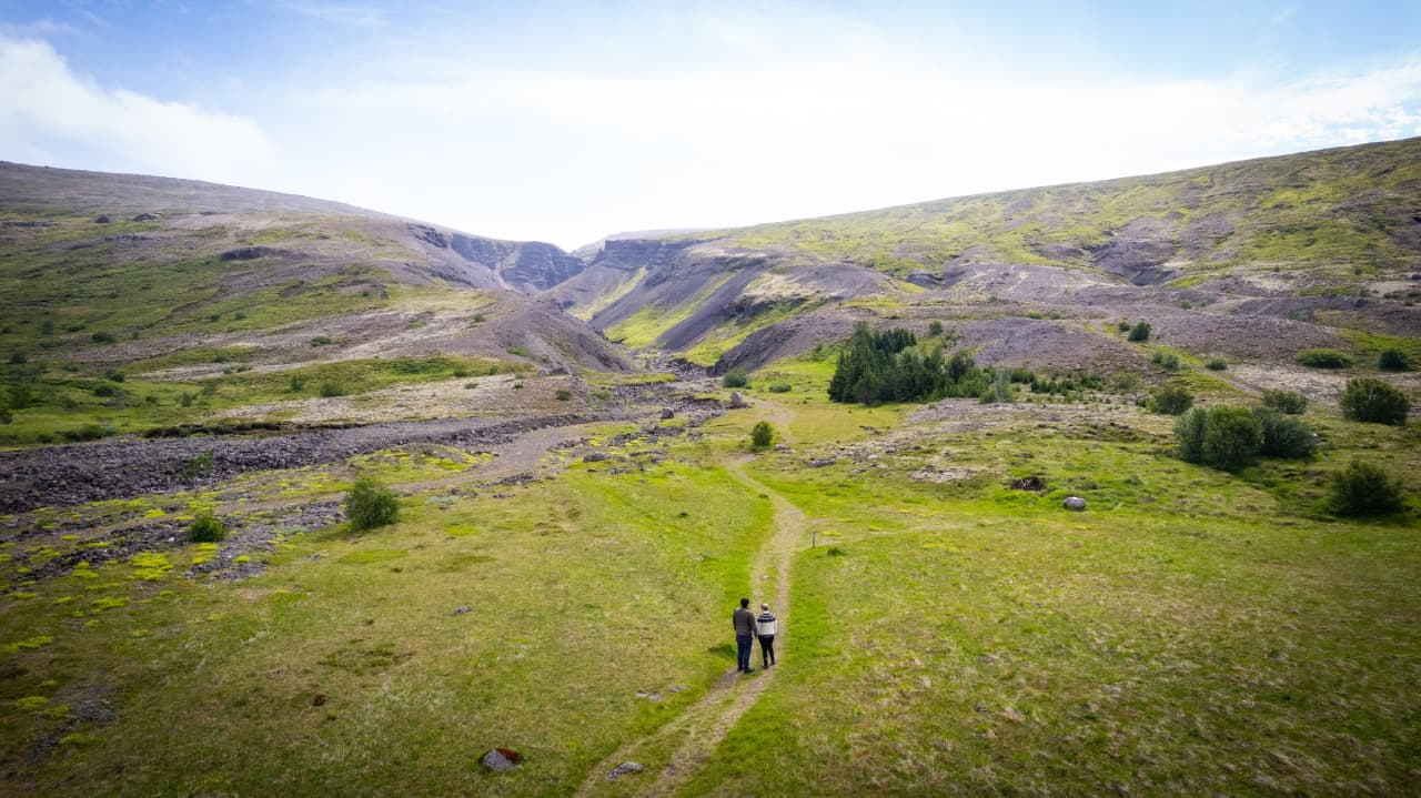 Two people walking a narrow path through open green and lava-colored hills near Húsafell.