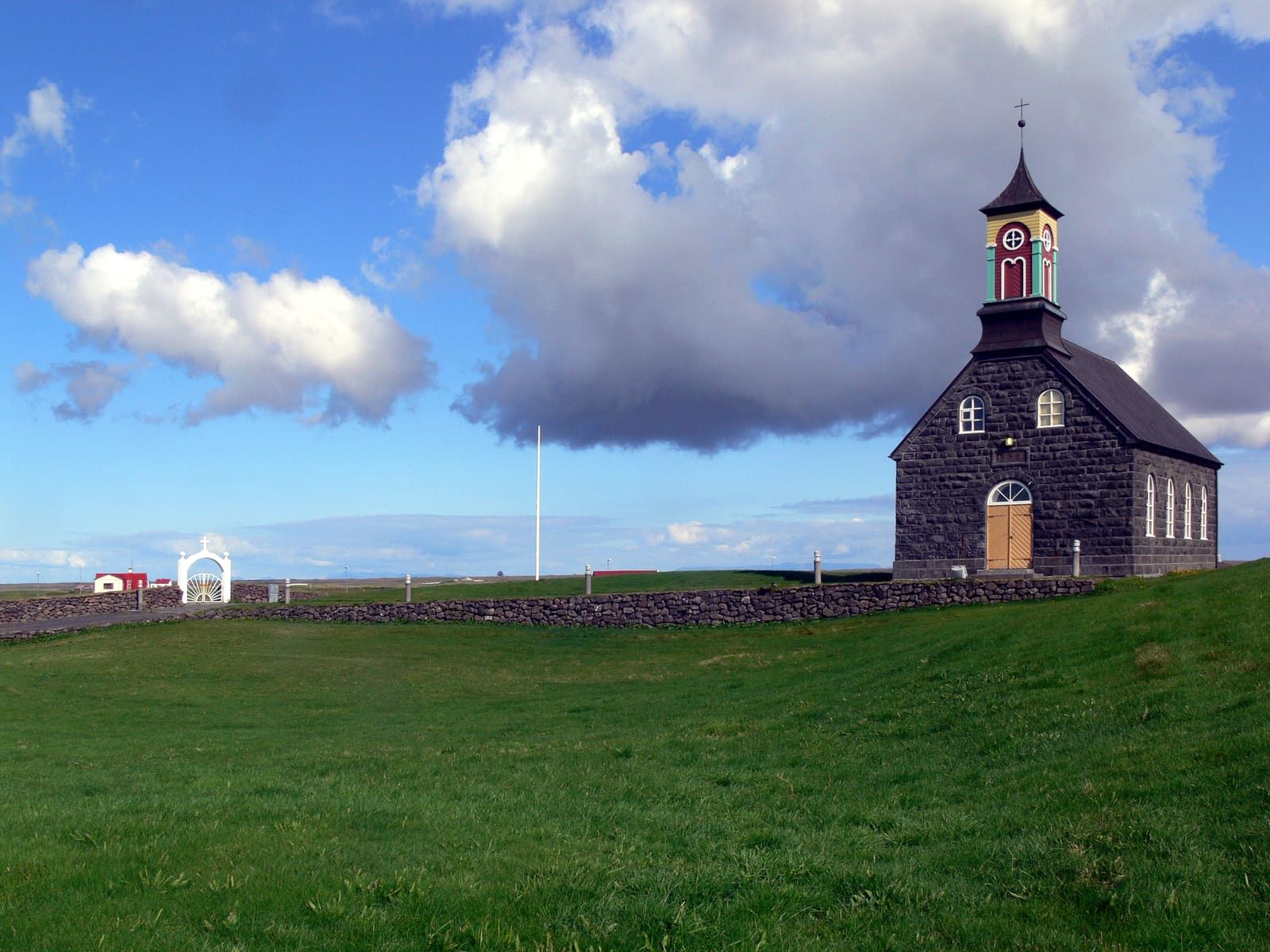 The entrance side of Hvalsneskirkja with dark stone walls, a red tower, and low Reykjanes coastline behind it.