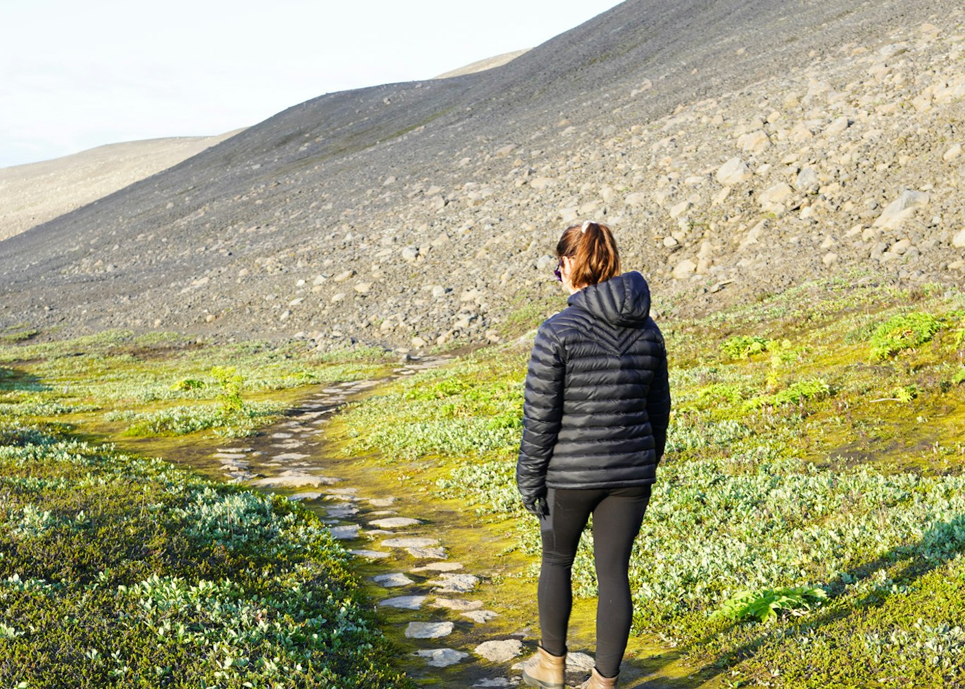 Visitor walking a stone path through low green vegetation at Hvannalindir below a bare highland slope.