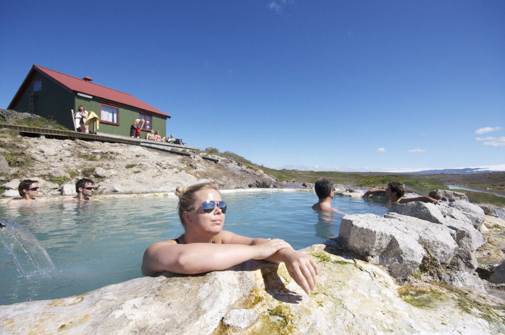 Travelers soaking in the Hveravellir hot pool beside a small green-roofed hut and distant Highlands views.