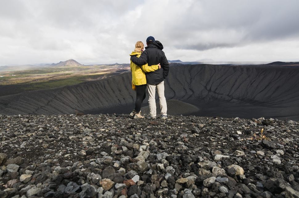 Two visitors standing on the Hverfell crater rim above the dark volcanic slopes.
