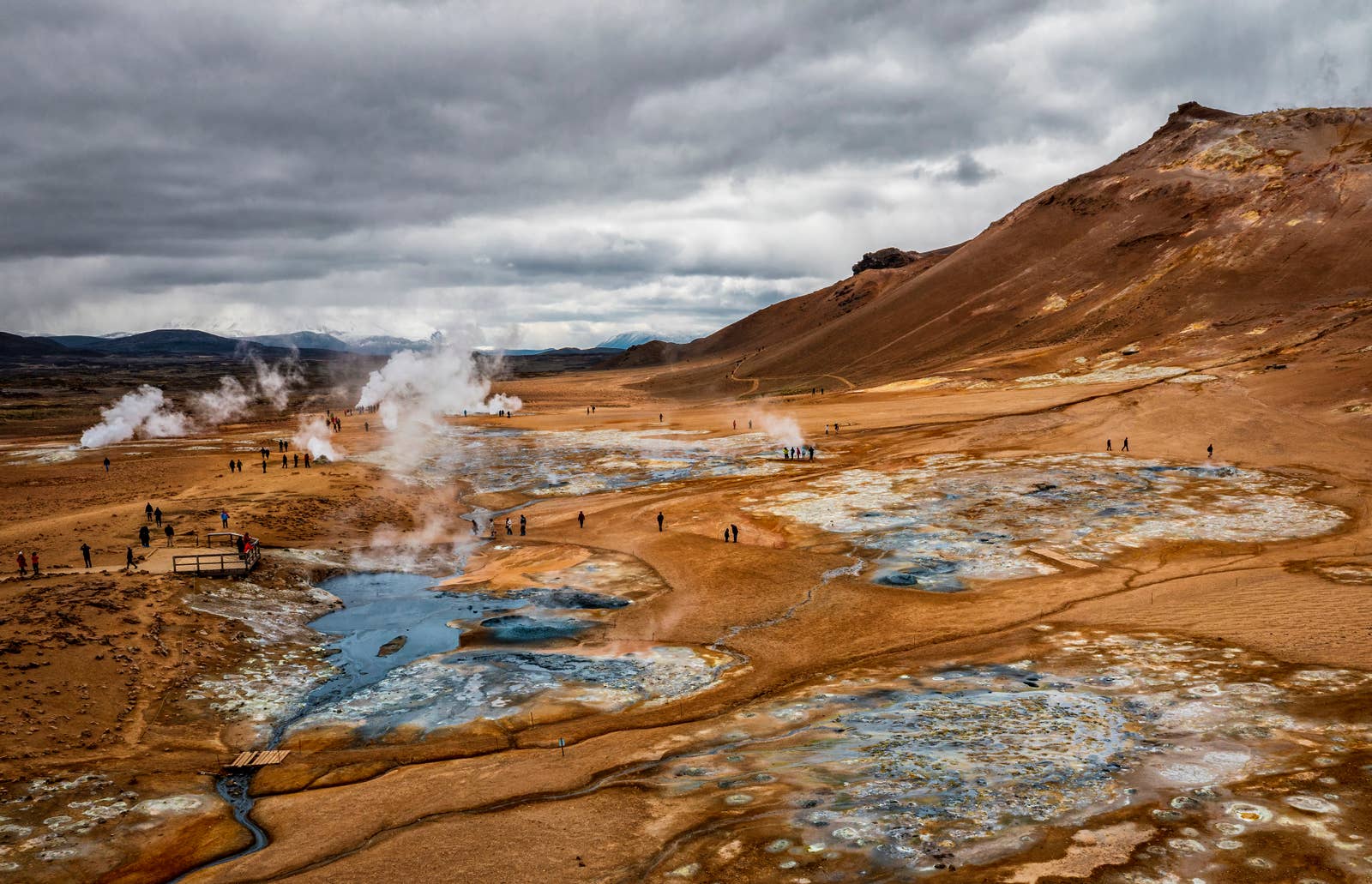 Wide view over Hverir Geothermal Area with steam plumes, mud pools, visitors, and the slopes of Námafjall.