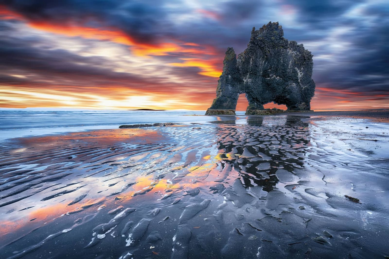Hvítserkur silhouetted against a colorful sunset over rippled wet sand.