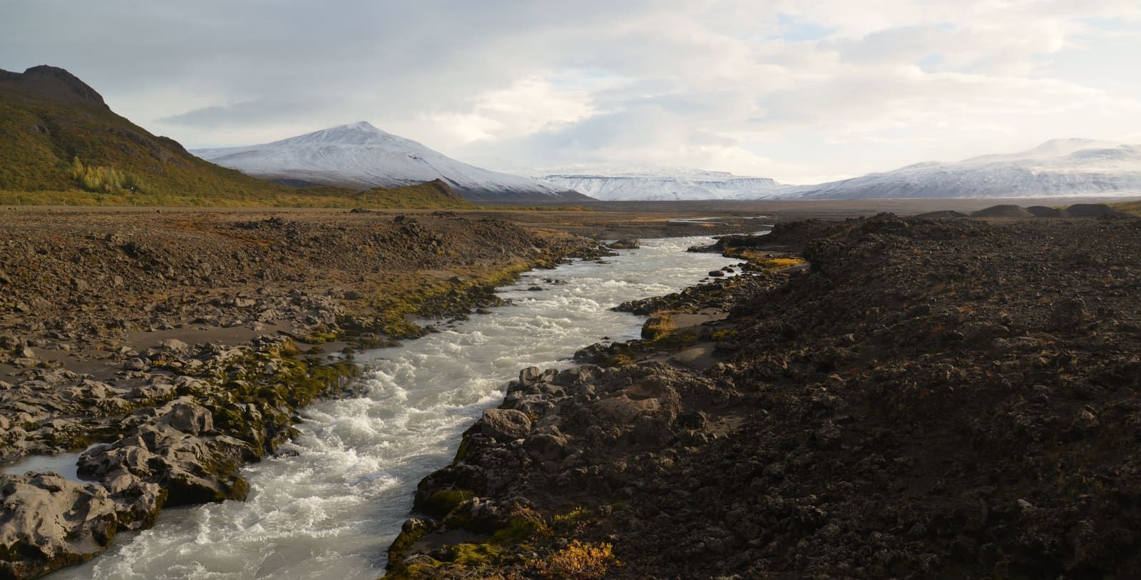 A fast river running through dark lava terrain at the foot of Kaldidalur with snow-dusted mountains beyond.