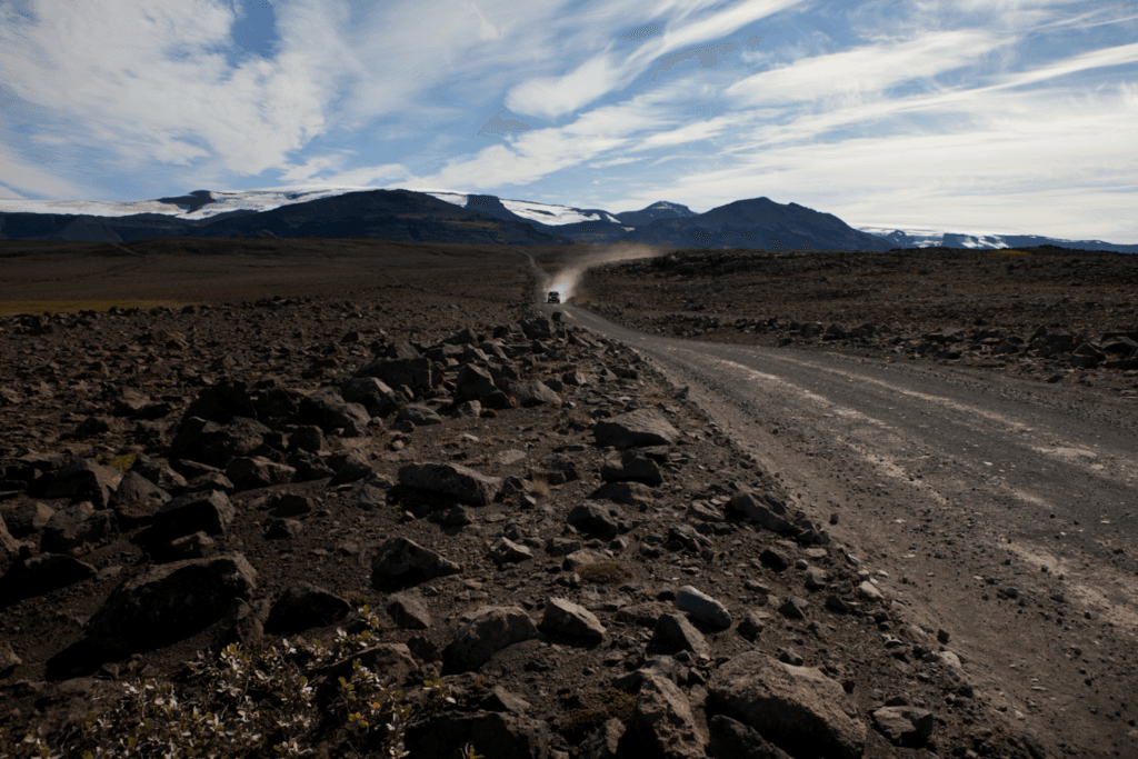 A gravel road crossing the rocky Kaldidalur valley with a vehicle dust trail and glacier views ahead.