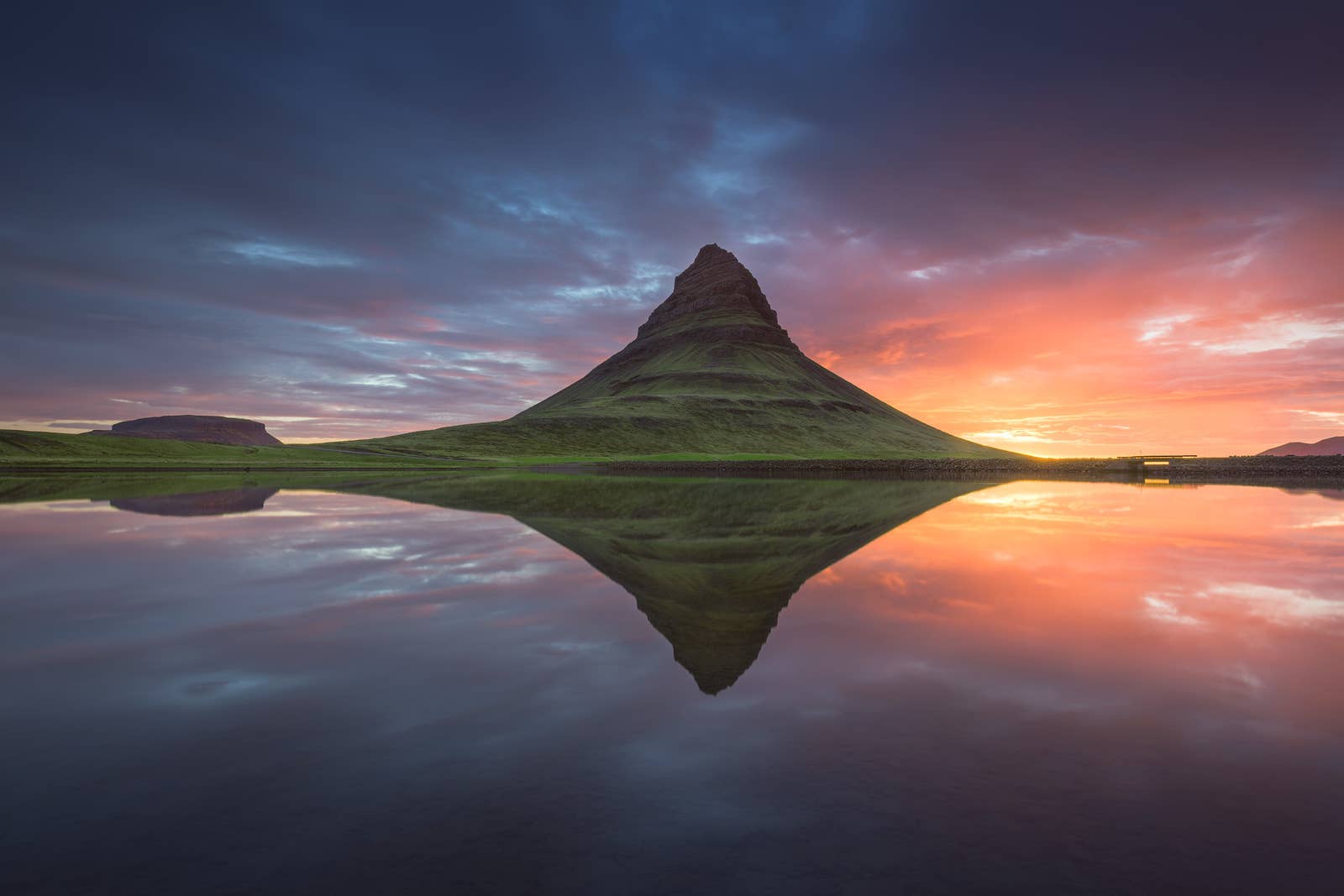 Kirkjufell reflected in calm water under colorful evening clouds.