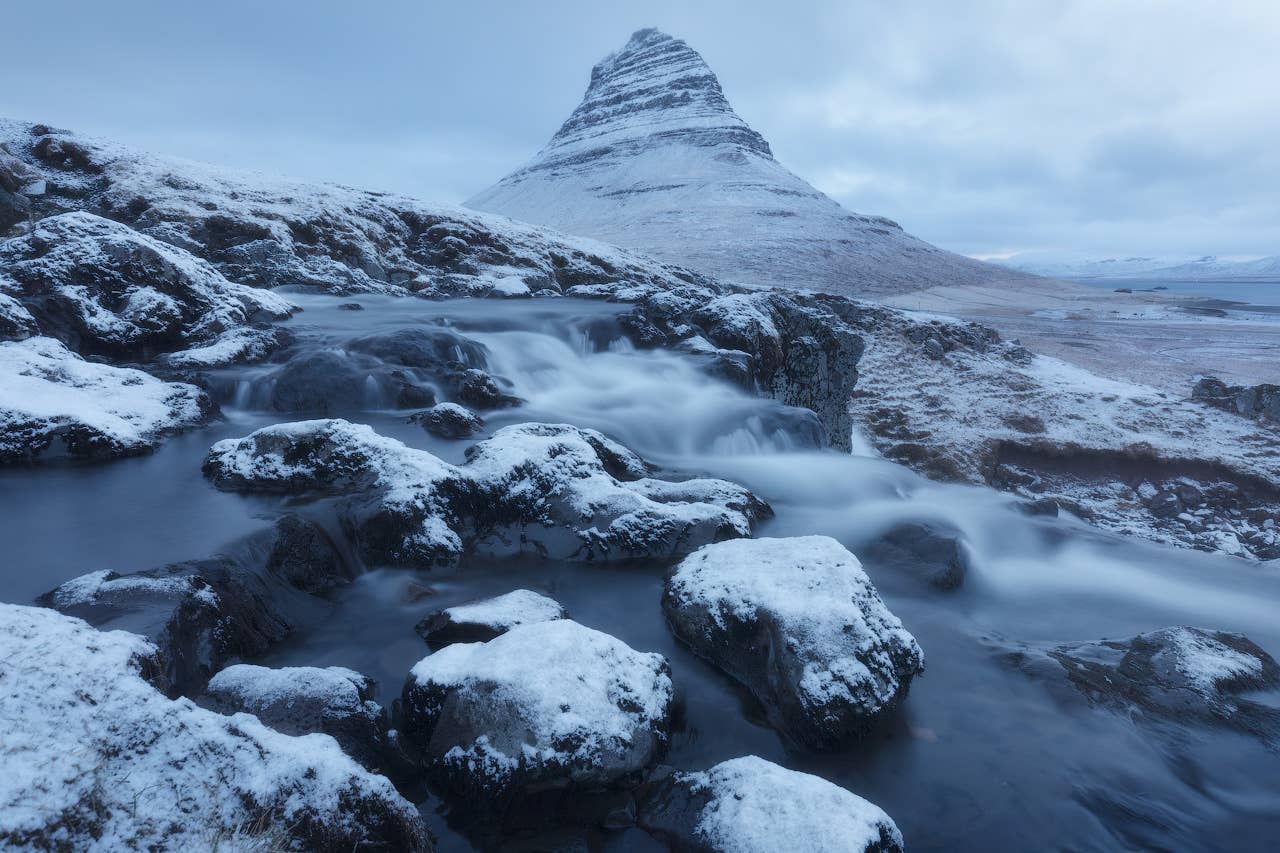 Snow-covered Kirkjufell behind the icy stream and rocks at Kirkjufellsfoss.