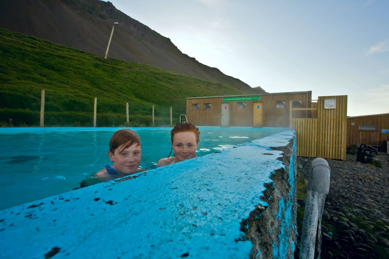 People in Krossneslaug beside the simple pool building and green hillside in Strandir.
