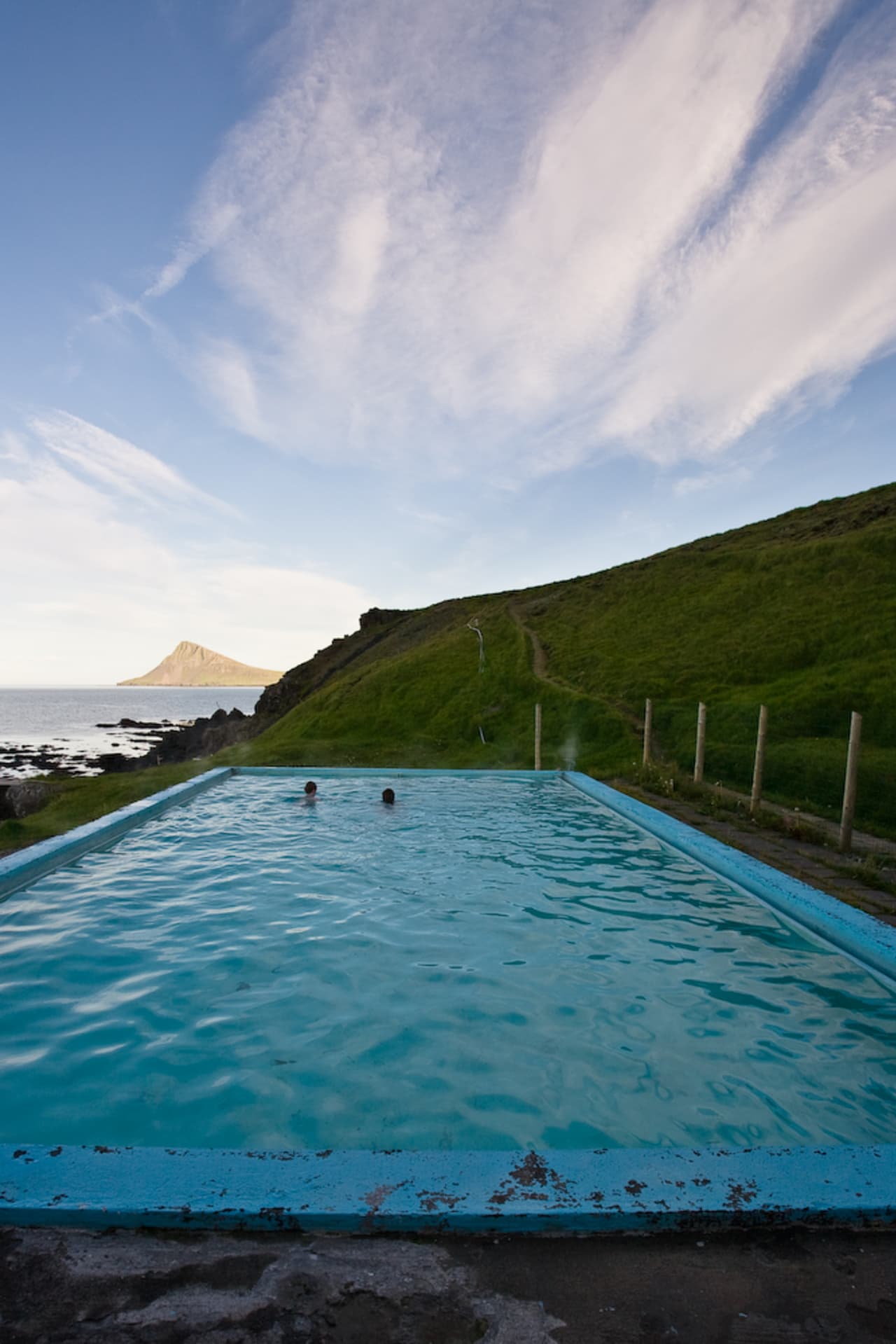 Krossneslaug pool looking toward the ocean, green hillside, and distant Strandir mountain.