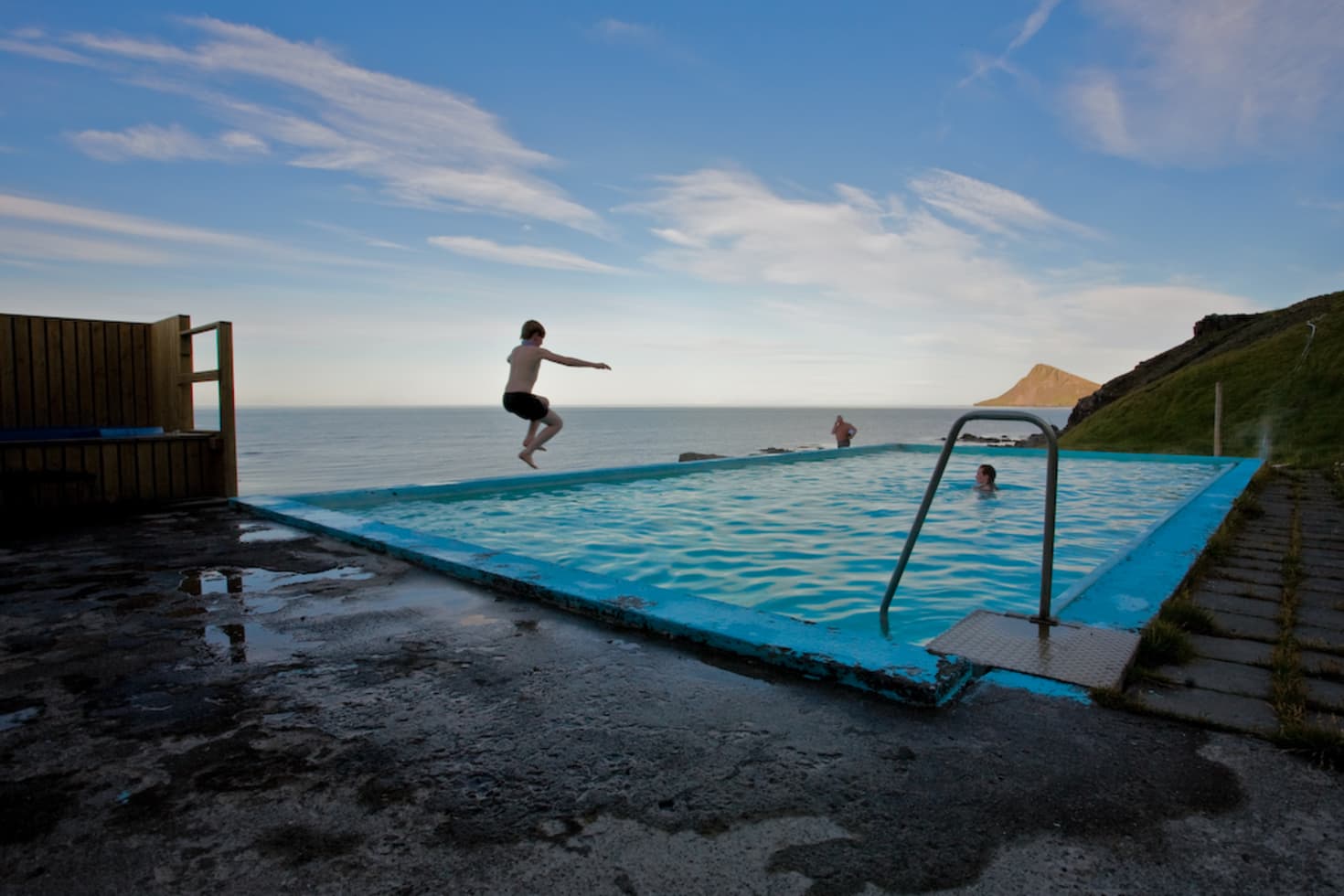 Swimmer jumping into Krossneslaug with the blue pool edge, open ocean, and green Strandir coast behind.