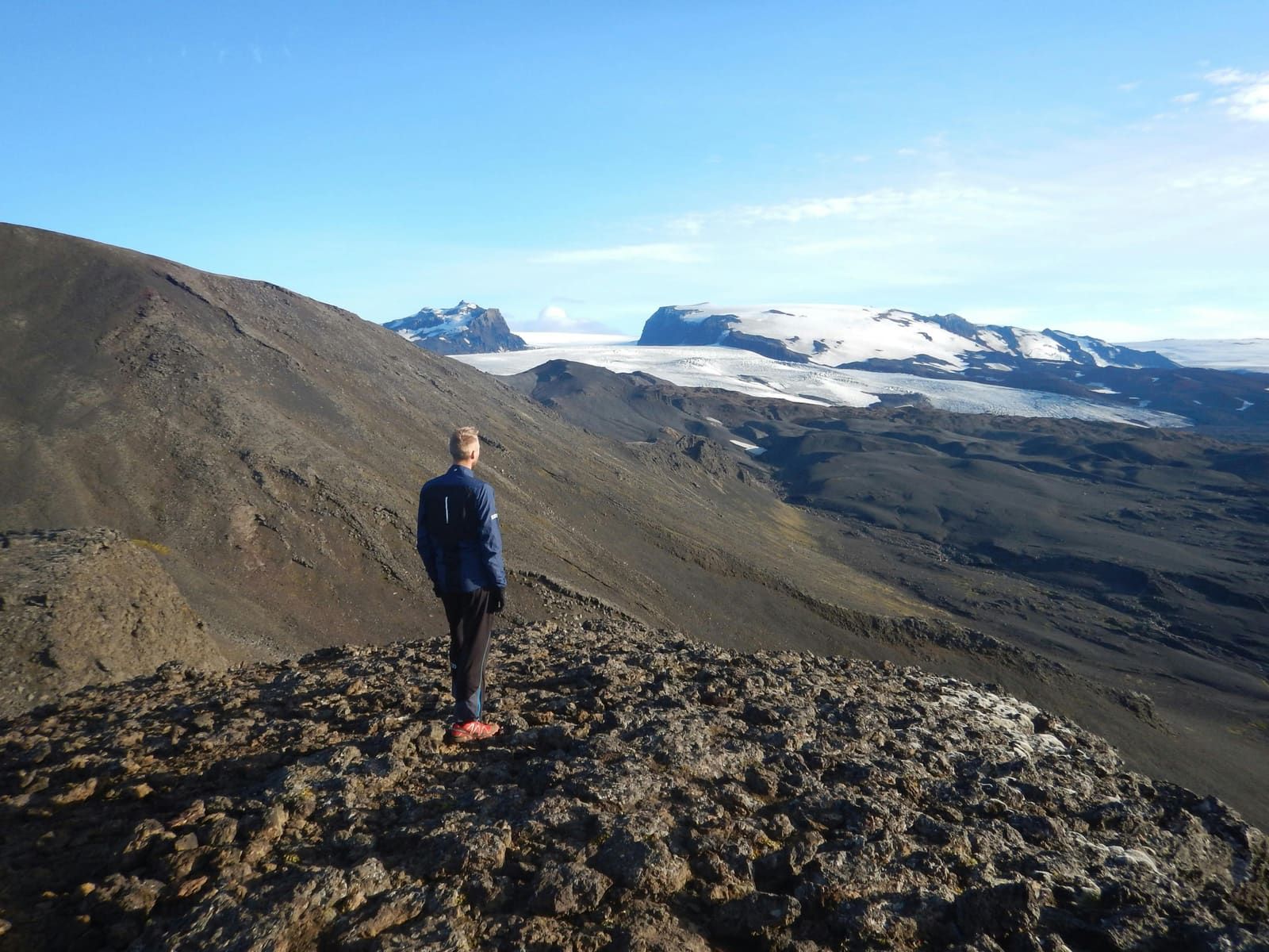 Hiker standing on dark rocky terrain above Kverkfjöll with glacier tongues and snow-covered mountains beyond.