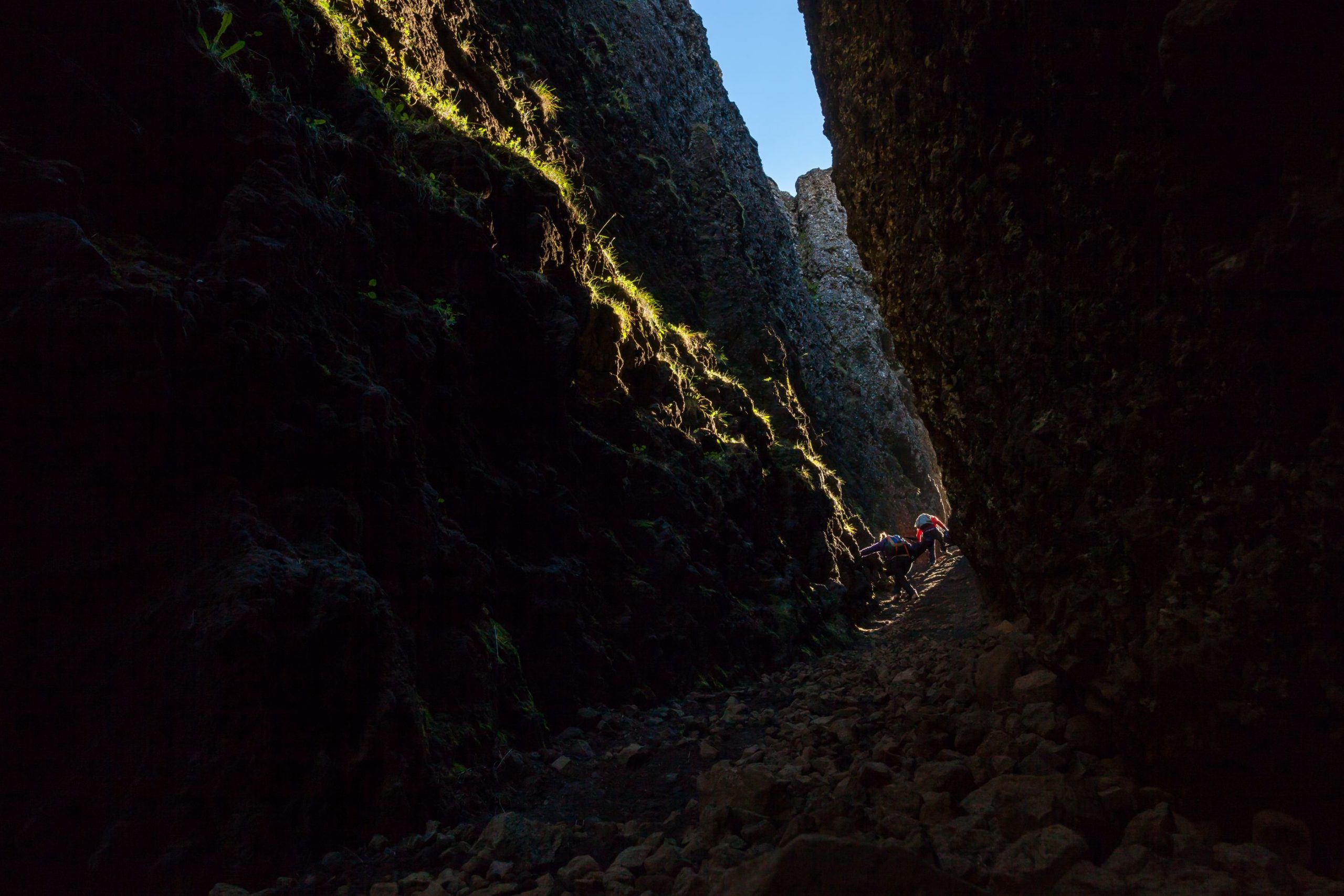 Hikers moving through the darker interior of Lambafellsgjá over rocky uneven ground.