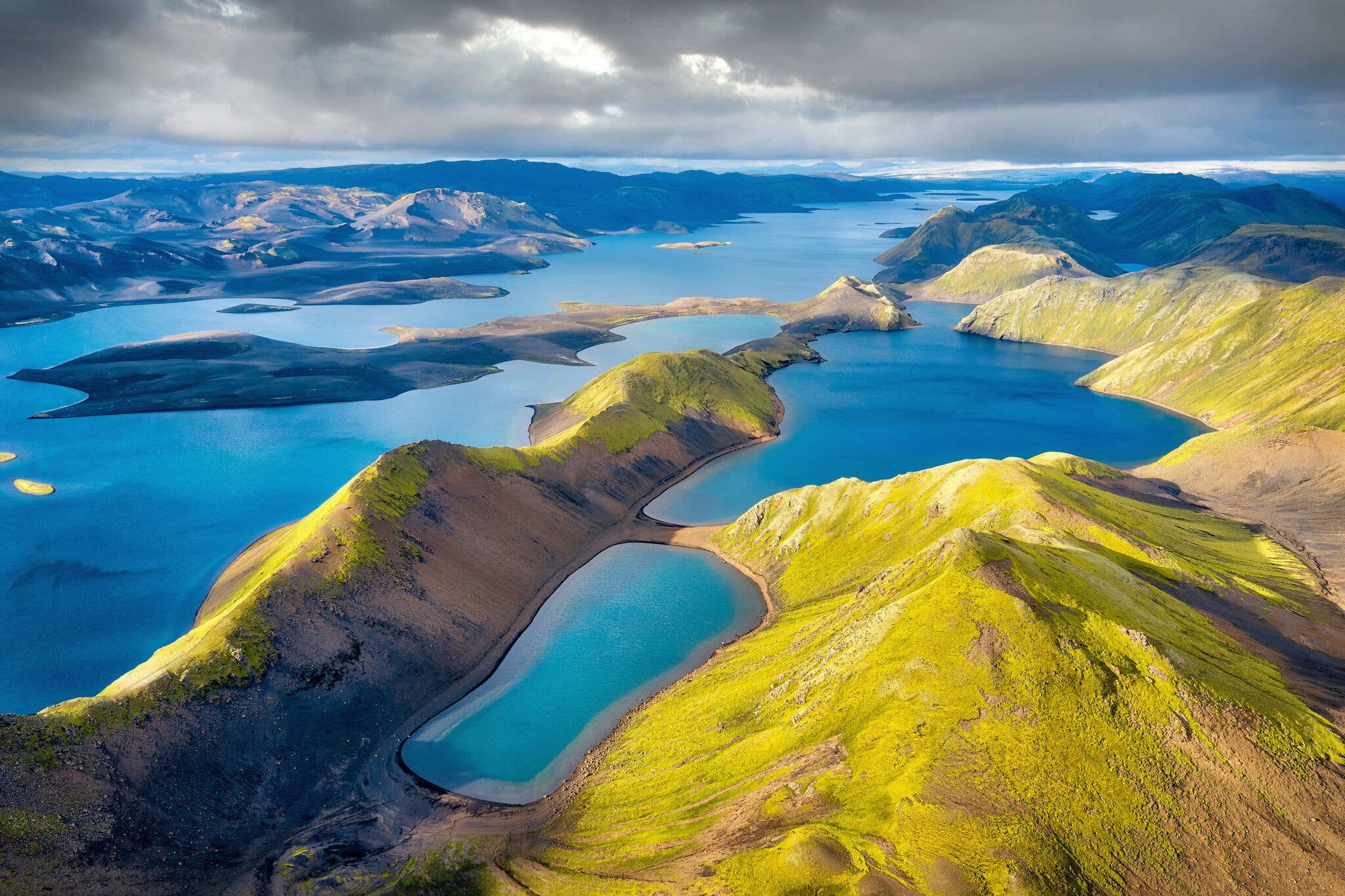 Aerial view of Langisjór with blue water channels, black sand, green ridges, and cloud shadow.