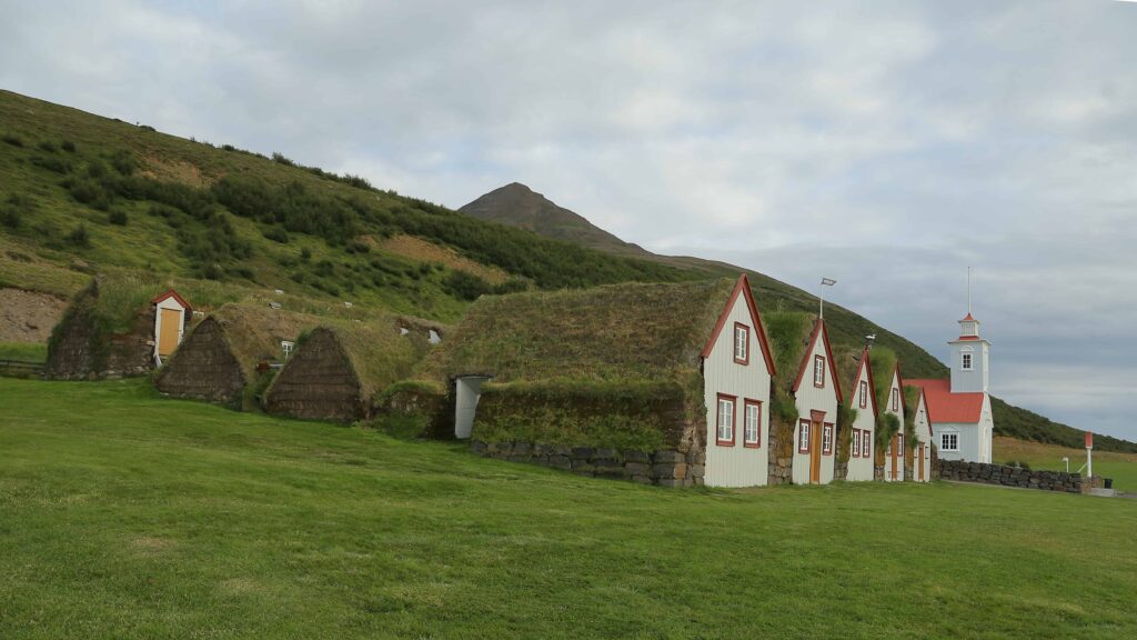 Wide farmyard view at Laufás with turf-roofed buildings, white gables, church, and green hillside.