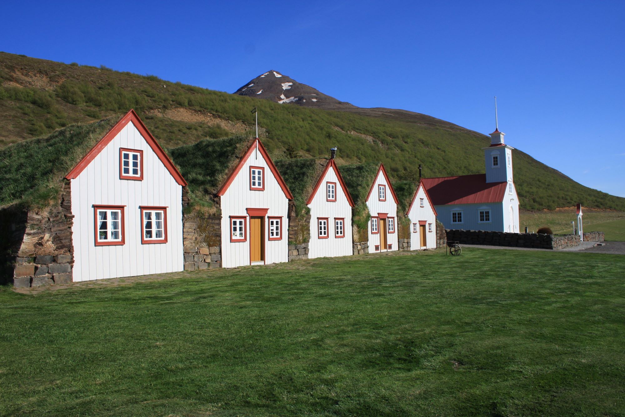 Laufás turf farm with white gables, turf roofs, church, green hillside, and mountain in Eyjafjörður.