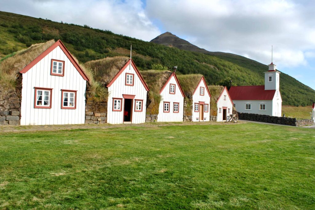 White timber gables and turf roofs at Laufás with the church and hillside behind.