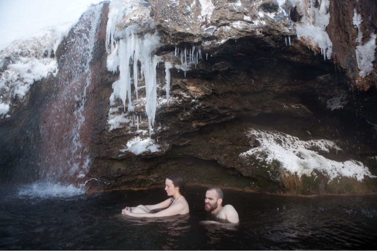 People soaking below the warm waterfall at Laugarvalladalur with ice on the rock wall.
