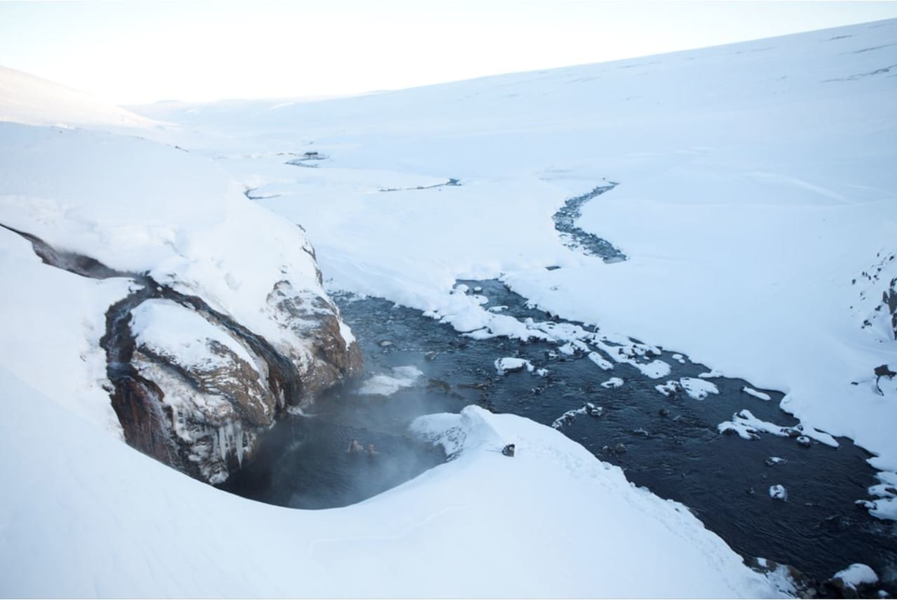 Snow-covered Laugarvalladalur valley with a dark river and steam rising near the hot waterfall.