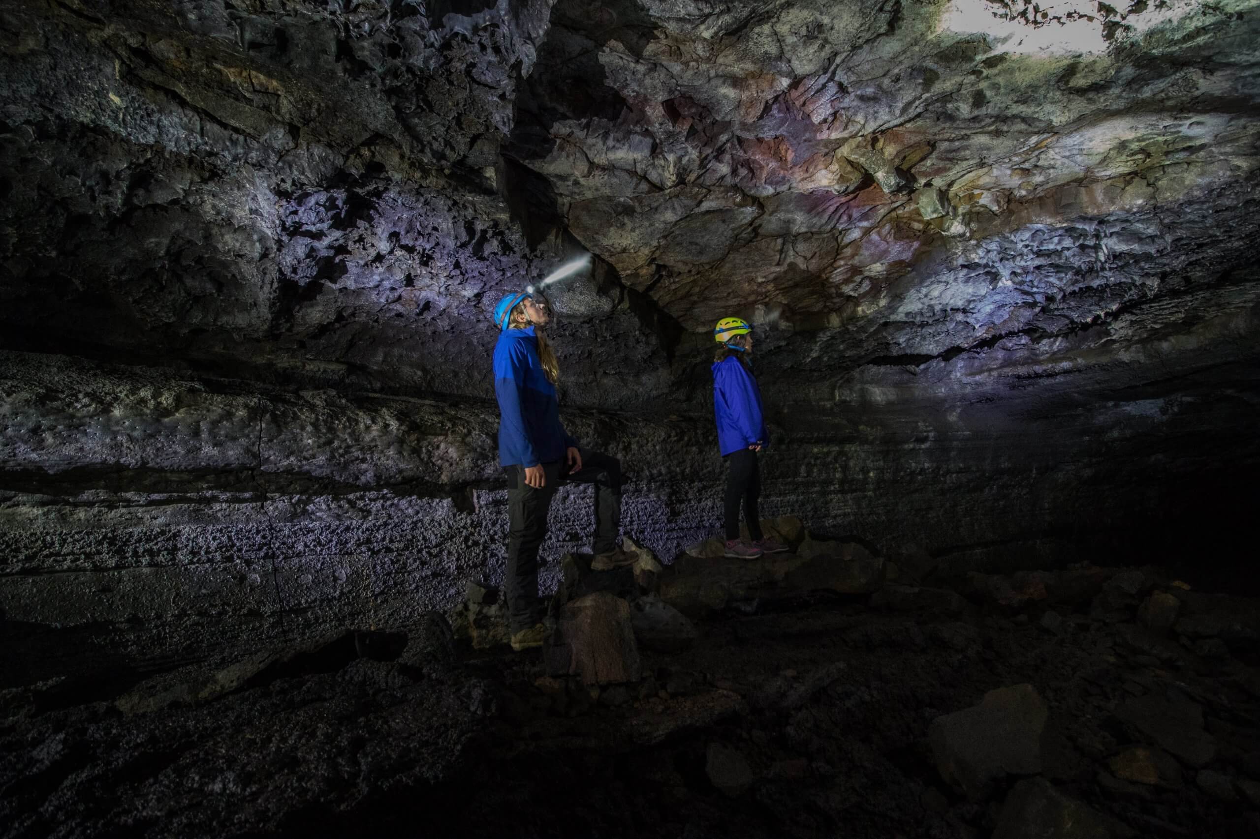 Two helmeted visitors using headlamps inside a broad dark chamber of Leiðarendi lava tube.