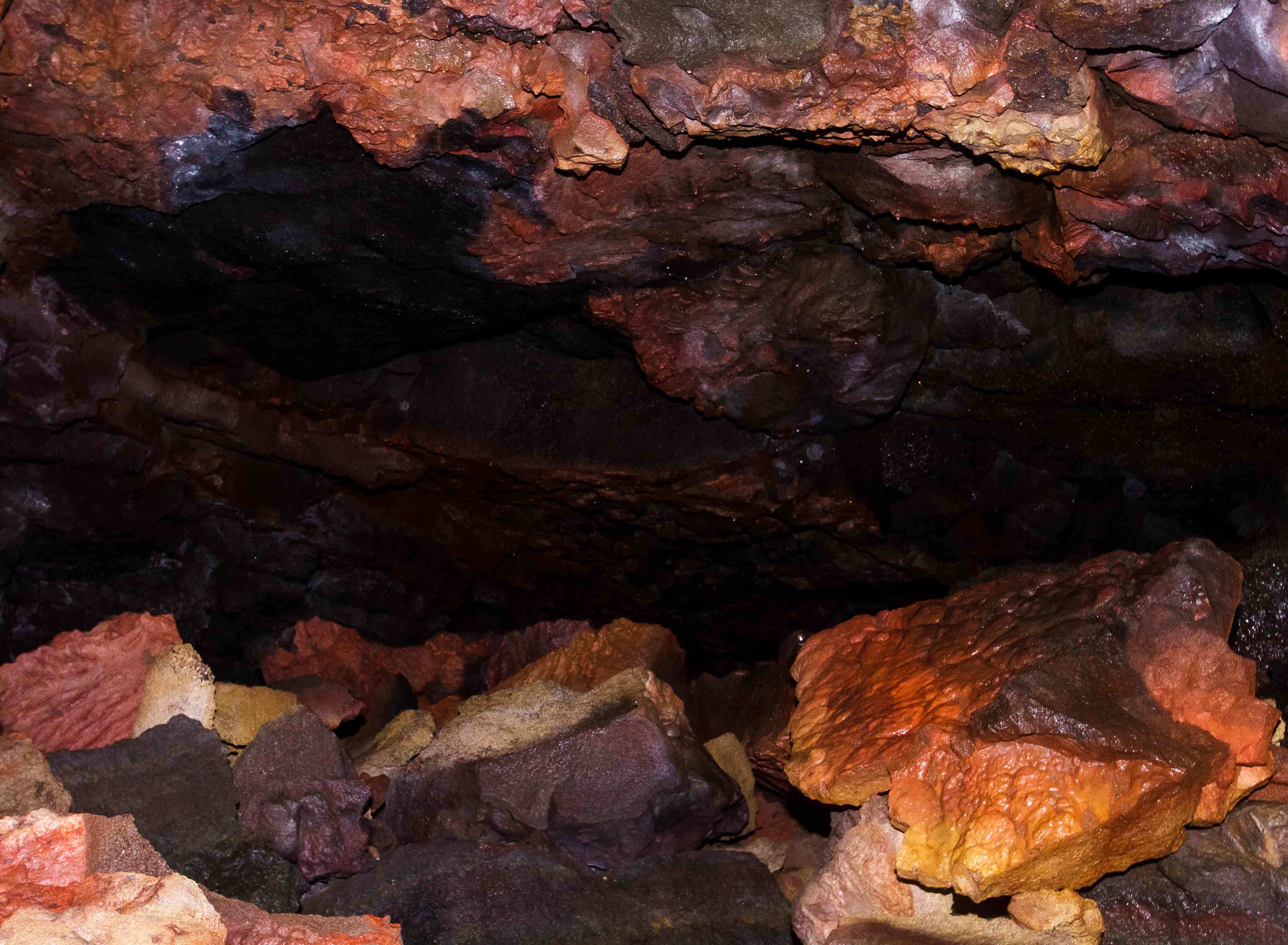 Red, orange, black, and pale mineral-stained lava rocks inside Leiðarendi Cave.