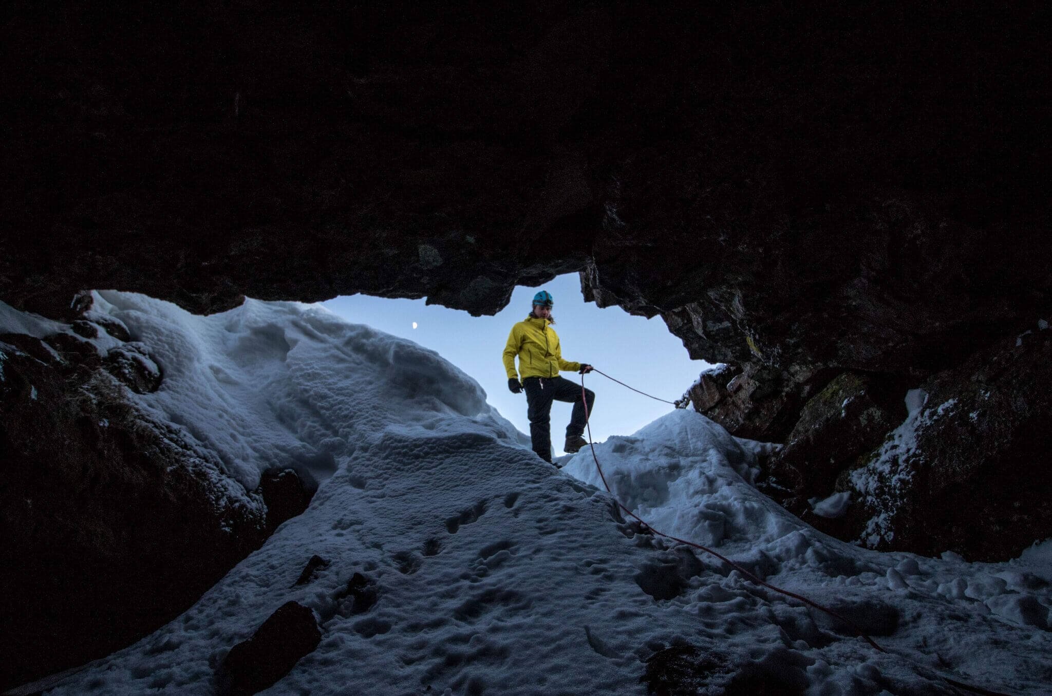 A helmeted guide standing by the snowy Leiðarendi cave entrance with a rope over uneven lava and ice.