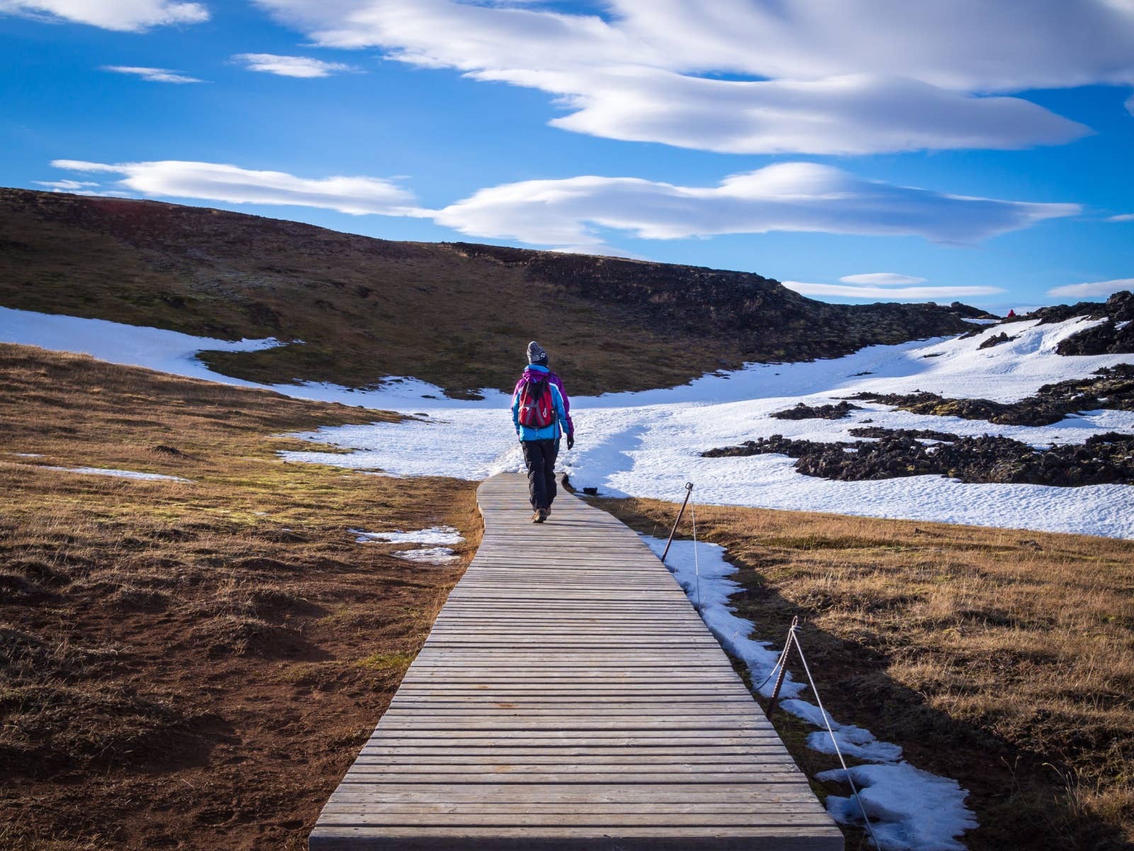 Wooden boardwalk leading through snow patches and dark lava toward Leirhnjúkur in the Krafla area.