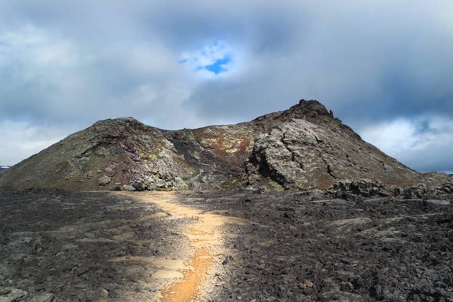 Narrow pale path crossing black lava toward the rocky Leirhnjúkur crater under cloudy sky.