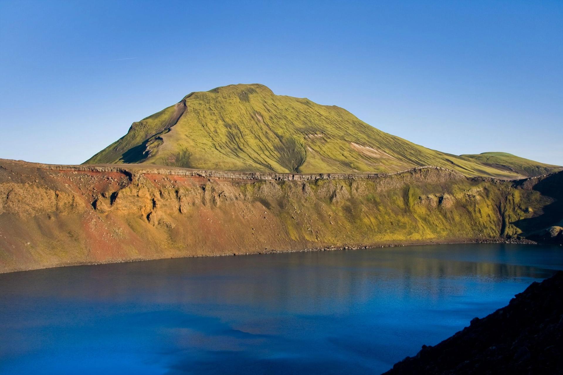 Blue water inside Ljótipollur crater below steep green, red, and brown volcanic slopes.