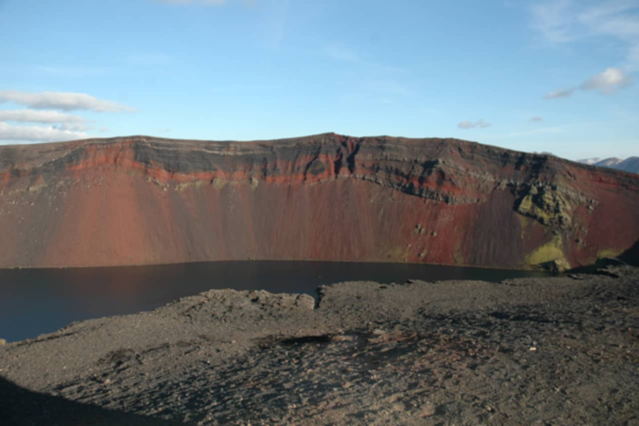 Red and black volcanic rim of Ljótipollur crater above a dark lake edge.