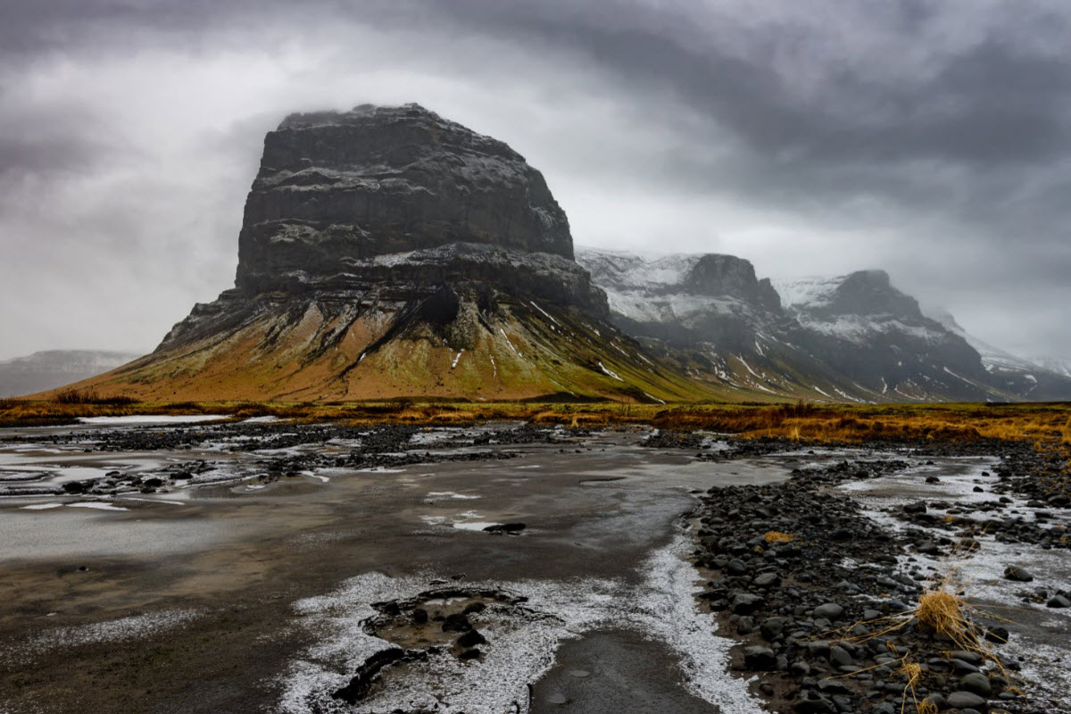 Storm clouds over Lómagnúpur with wet black stones and low vegetation in the foreground.