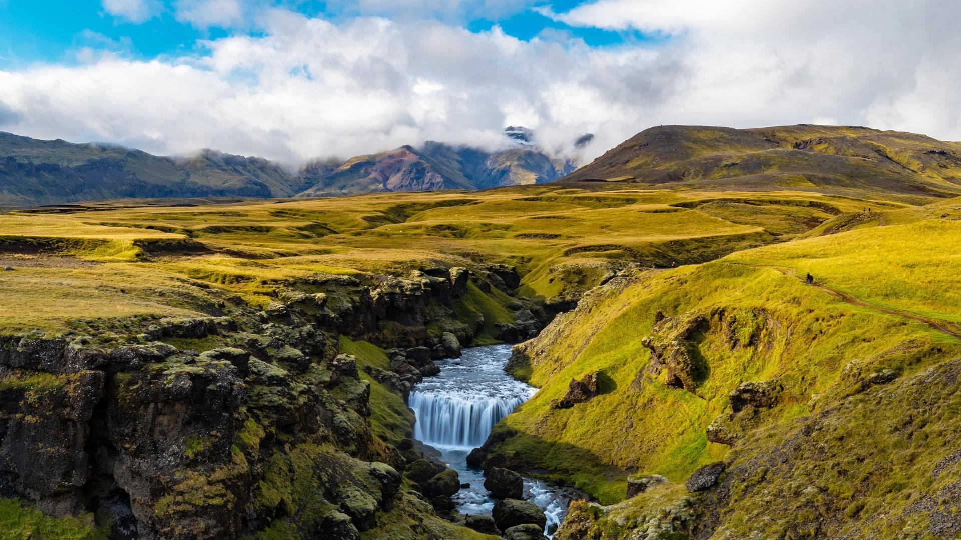 Green Fimmvörðuháls approach terrain with a small waterfall and mountains under cloud.