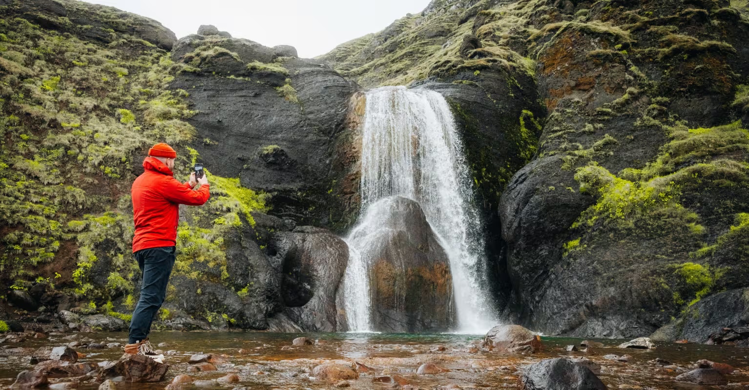 A visitor photographing Helgufoss waterfall in a rocky ravine near Mosfellsdalur.