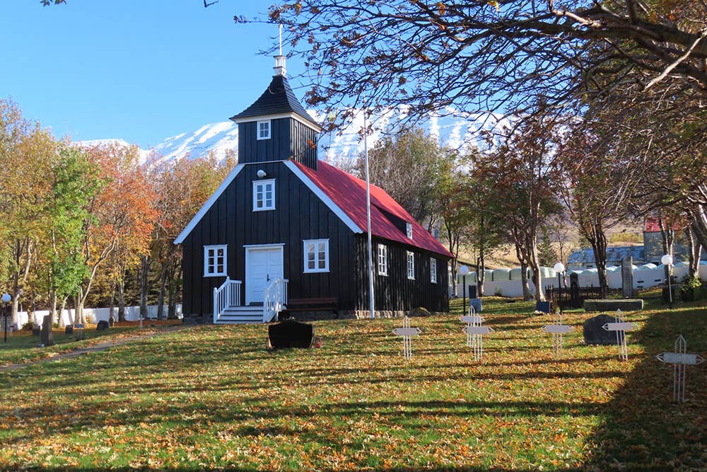 Munkaþverárkirkja with dark timber walls, red roof, churchyard crosses, and mountains behind.