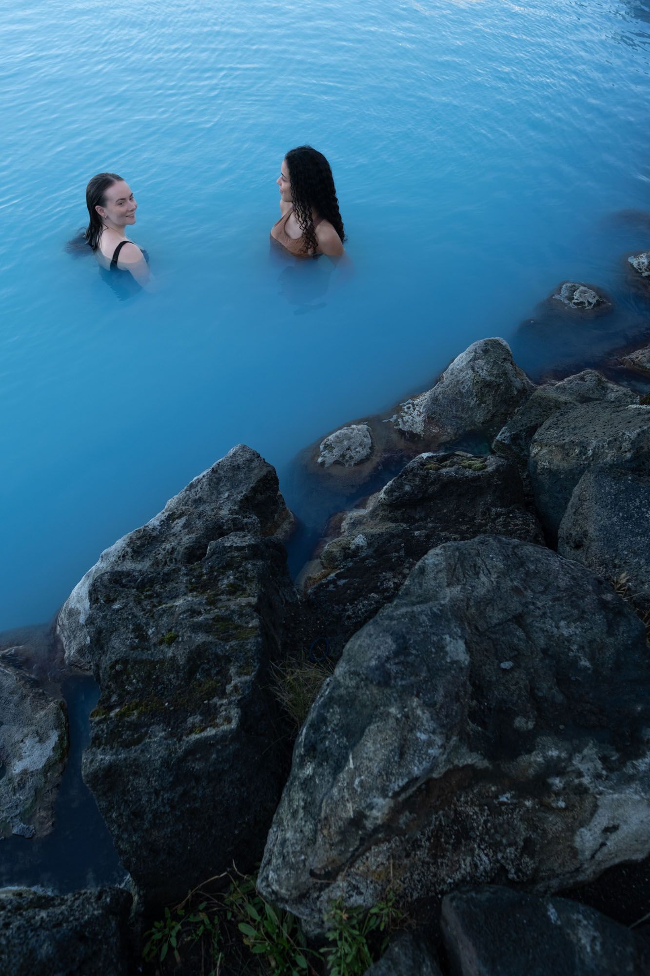 Two bathers in the blue geothermal water at Earth Lagoon Mývatn beside dark volcanic rocks.