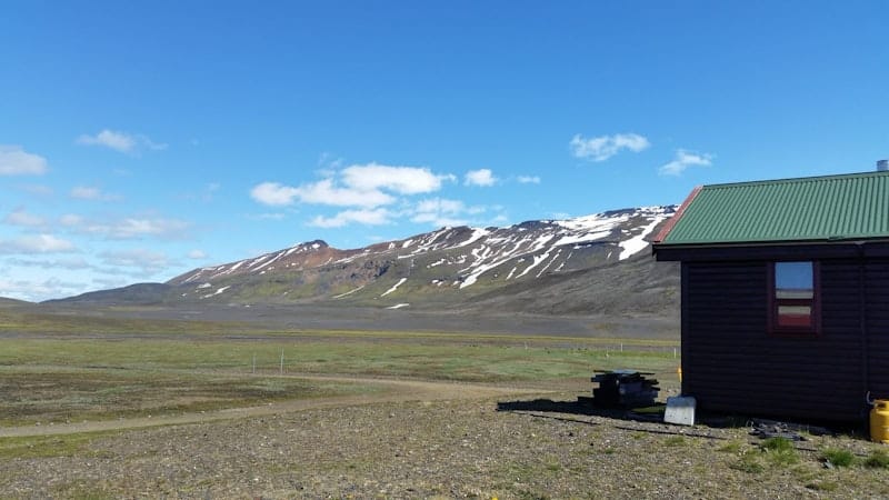 Nýidalur hut beside open Highlands ground and snow-streaked mountains in clear weather.