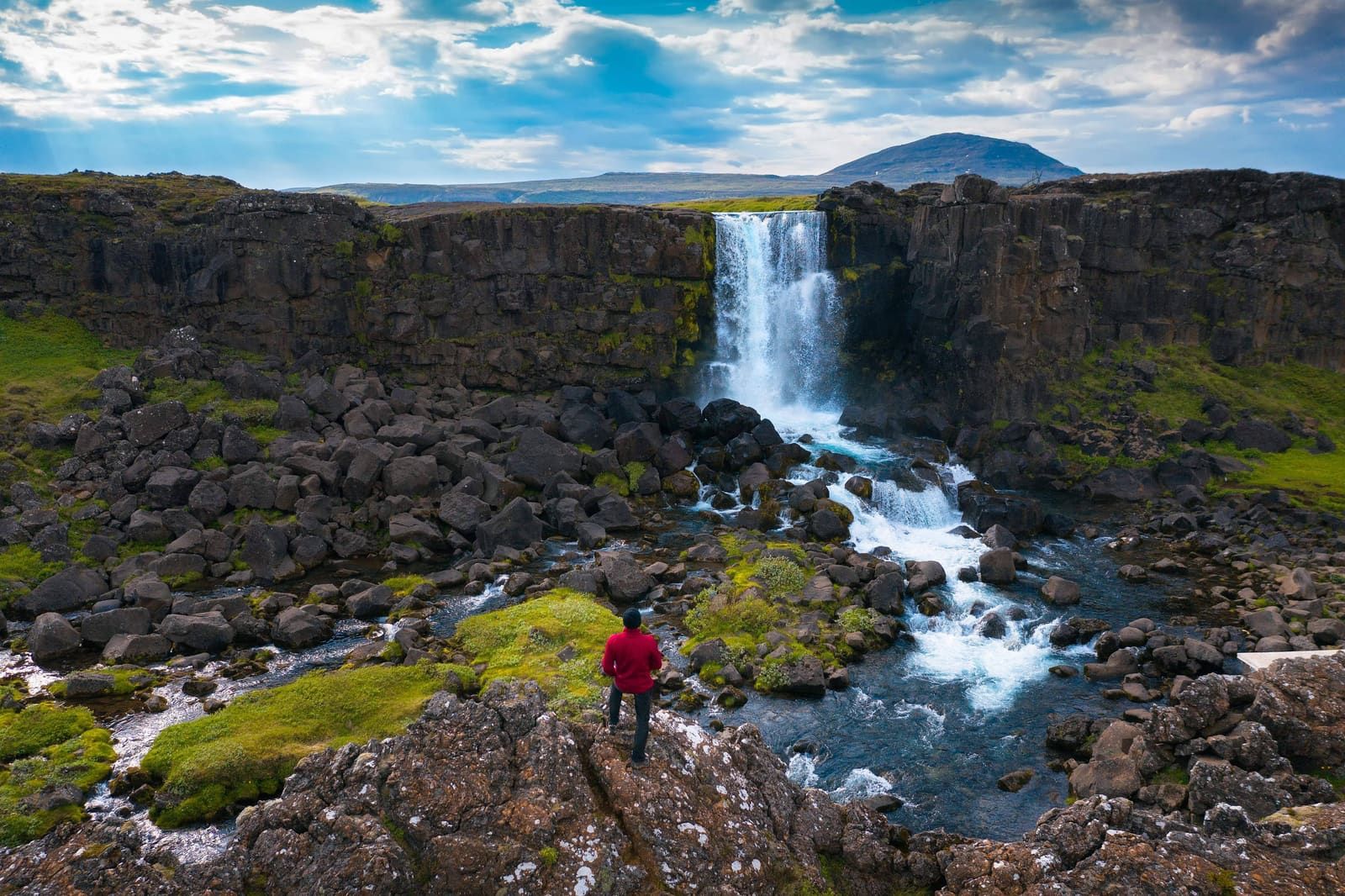 Visitor in a red jacket looking toward Öxarárfoss from mossy rocks beside the river.