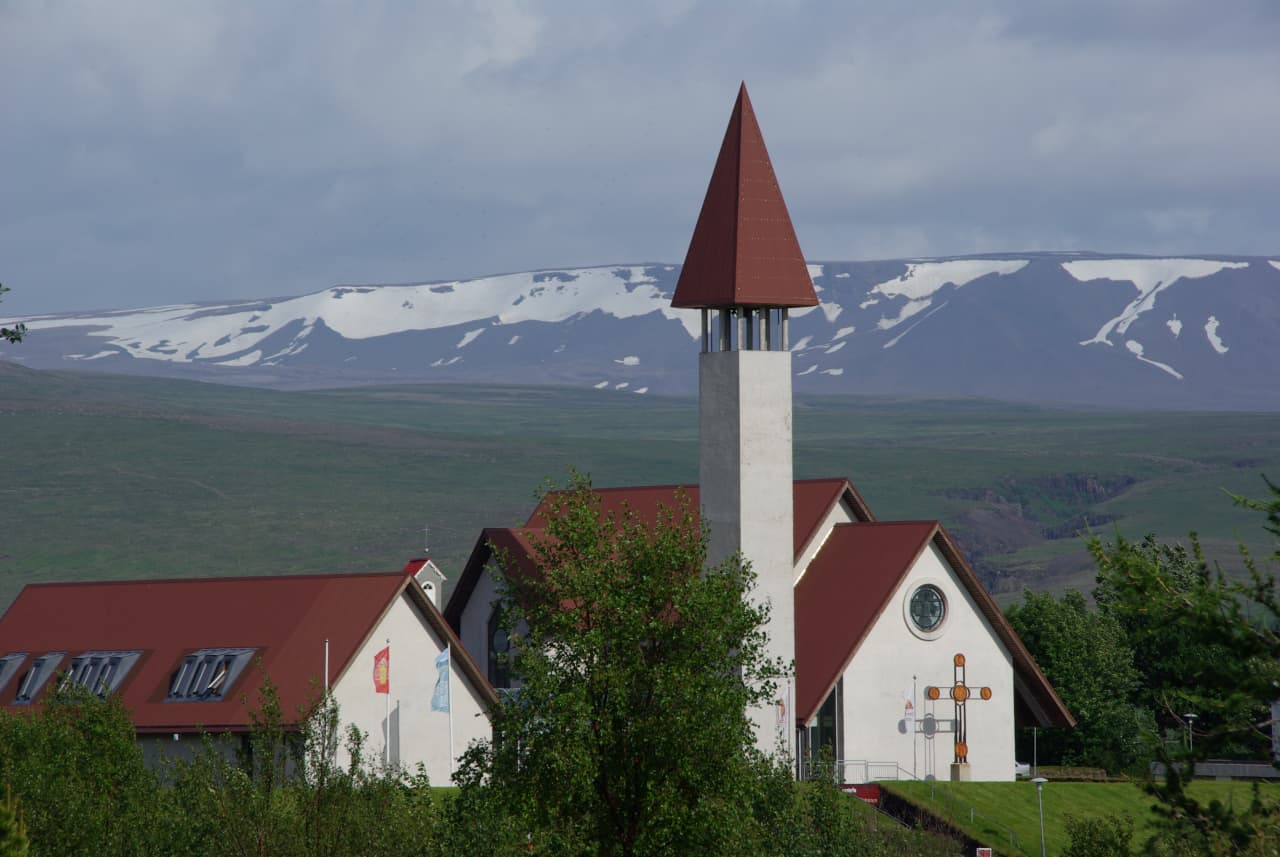 Reykholt church tower and red roofs below green hills with snow patches