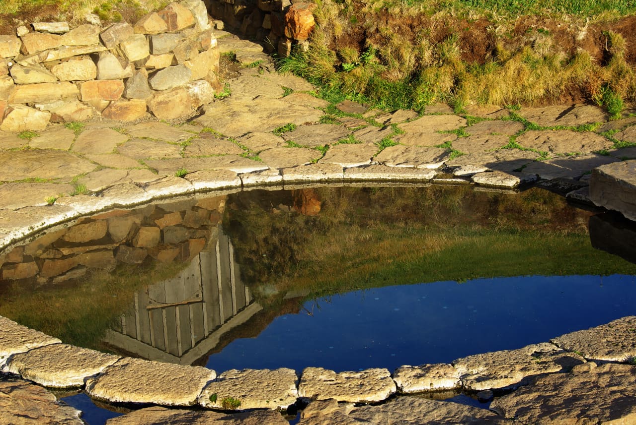 Stone edge and still water of Snorralaug reflecting the small turf-covered passage house