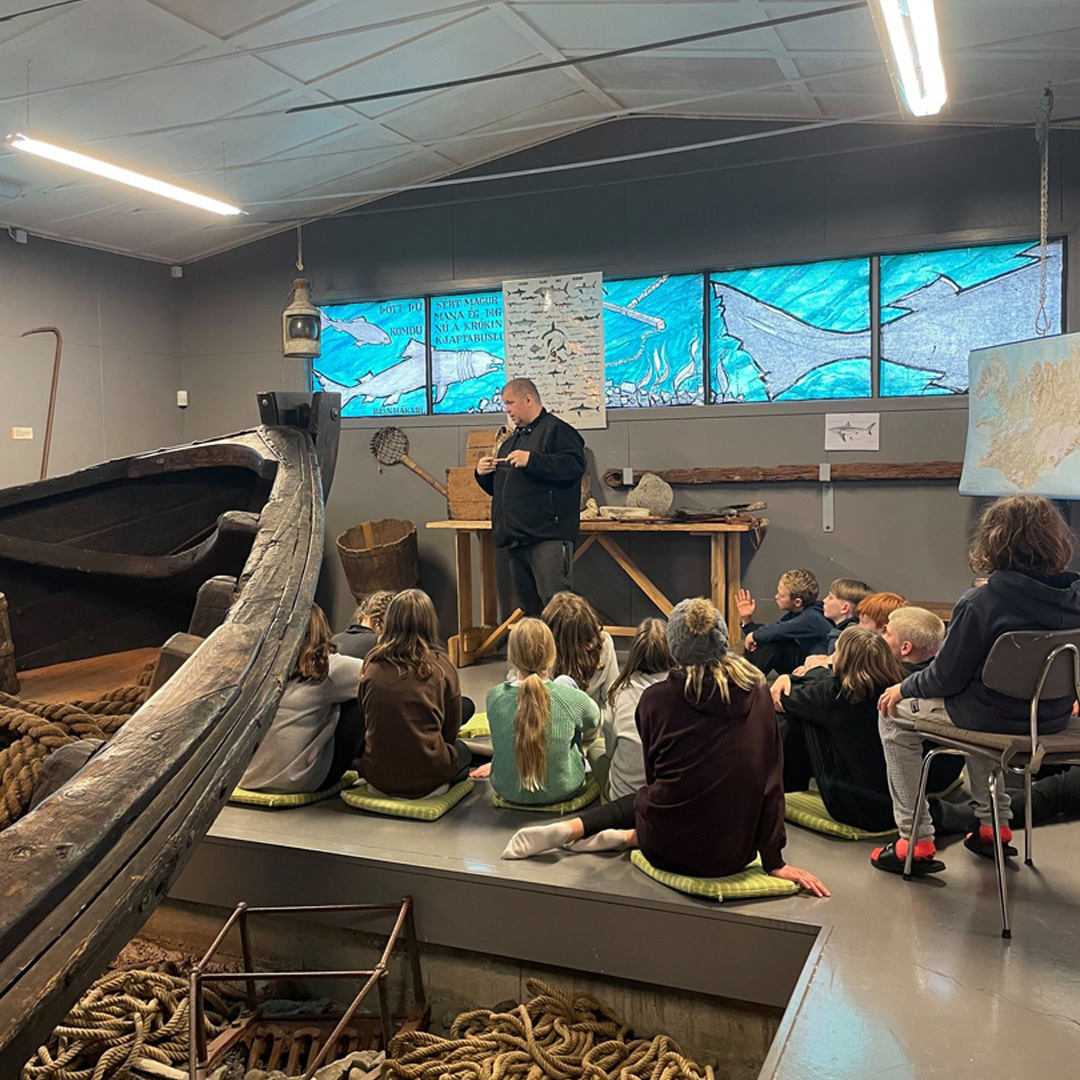 A museum guide speaking beside the Ófeigur shark-fishing boat inside Reykir Regional Museum.