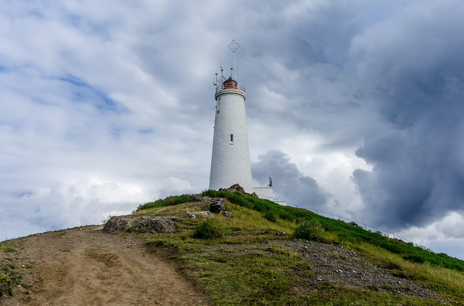 Close view of Reykjanesviti Lighthouse above a rough path and grassy volcanic hill under heavy clouds.