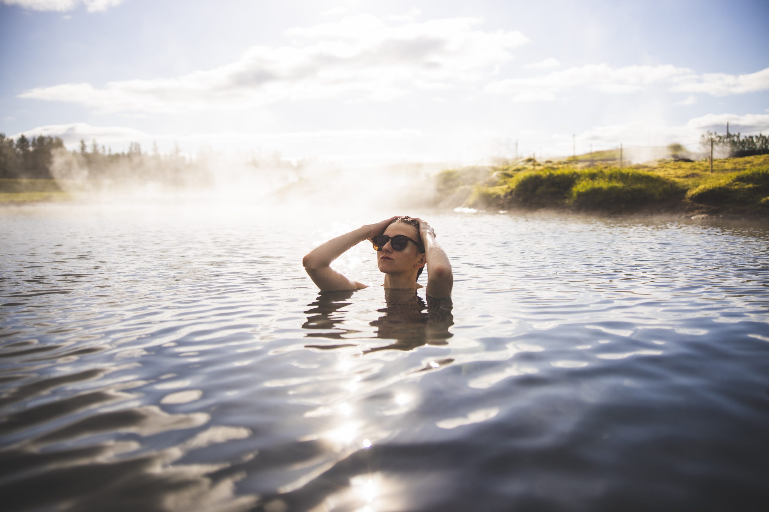 Visitor soaking in Secret Lagoon with low steam and open countryside behind the pool.