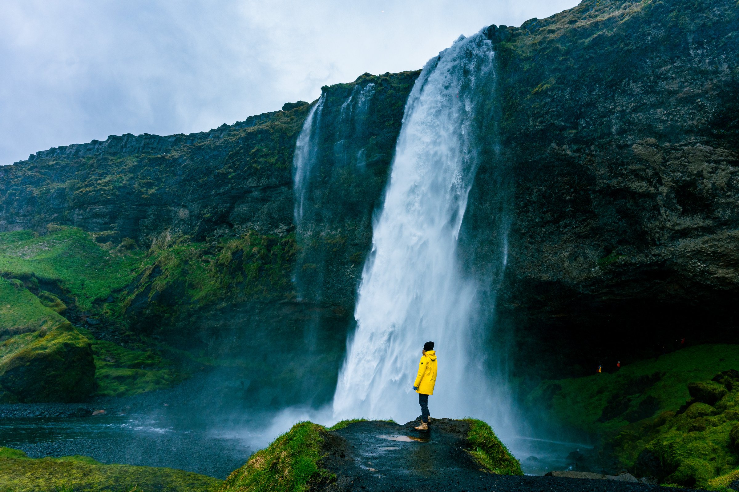 A visitor in a yellow jacket stands near the spray at the base of Seljalandsfoss.