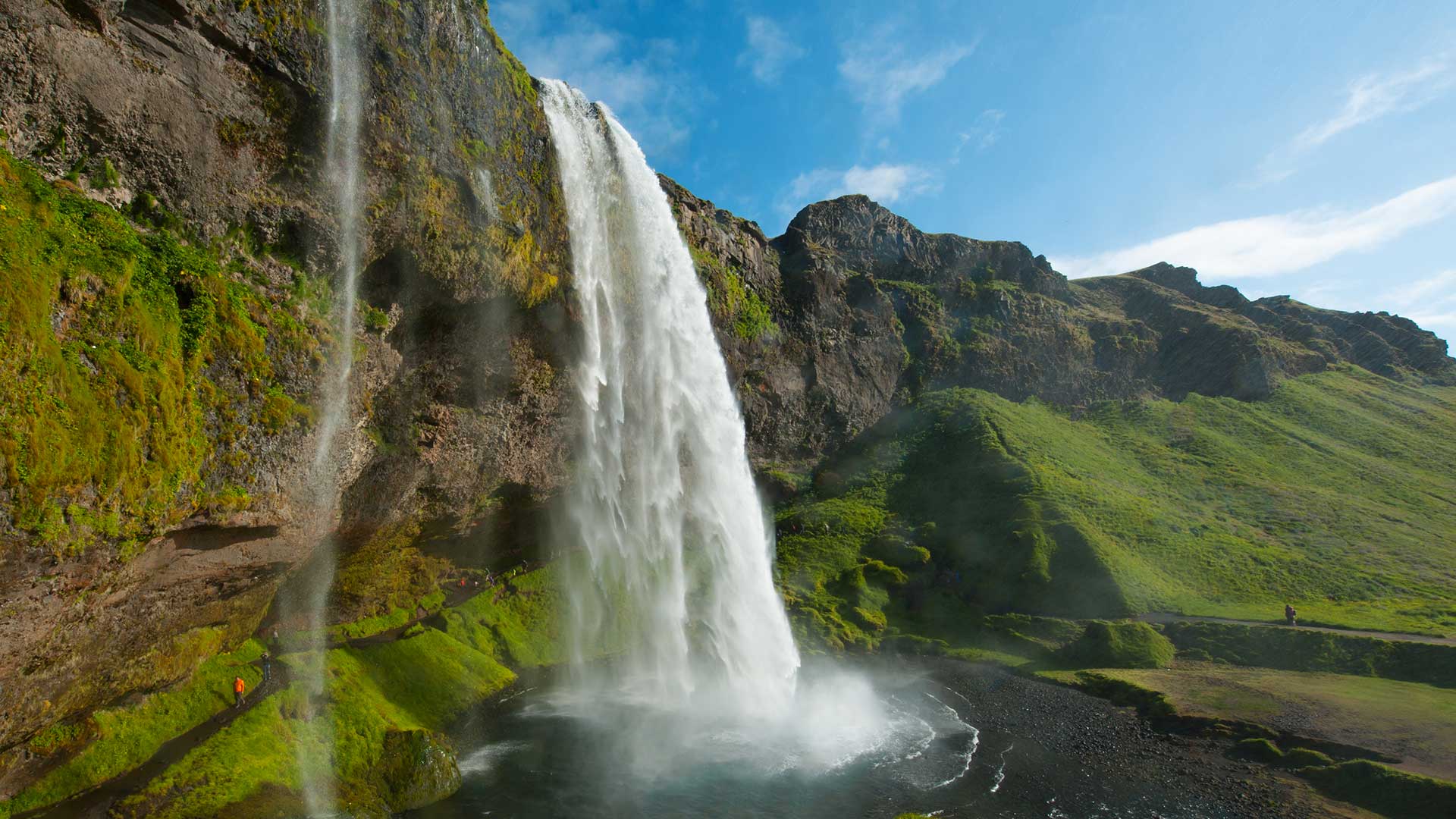 Summer view of Seljalandsfoss with the side path and green cliffs visible.