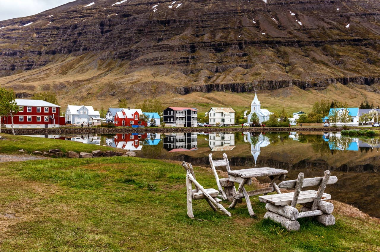 Colorful houses and Seyðisfjörður Church reflected in calm water below steep Eastfjords mountains.