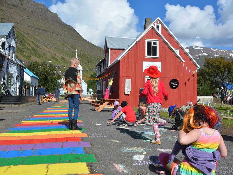 People on Rainbow Street in Seyðisfjörður with colorful painted road stripes, old houses, and steep mountains behind.