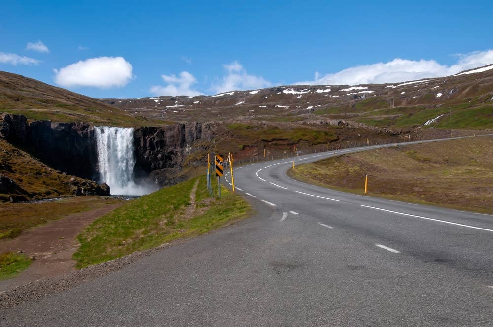 Gufufoss waterfall beside Route 93 near Seyðisfjörður with a curving road through open mountain terrain.