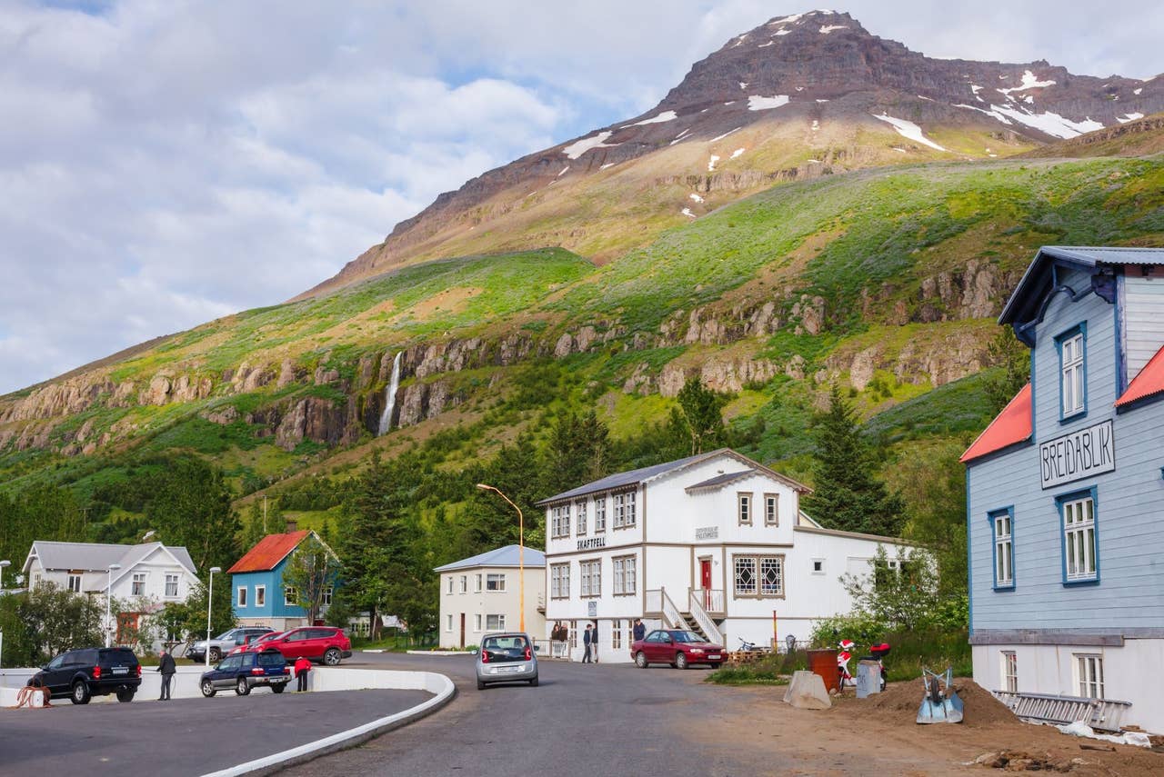 Street in Seyðisfjörður with old buildings, parked cars, waterfalls, and steep green mountains above town.