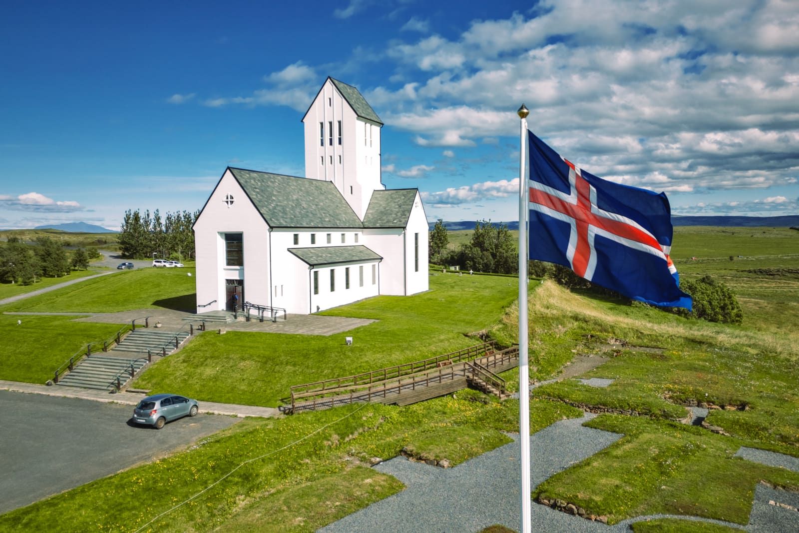 Skálholt Cathedral beside an Icelandic flag, with paths and low South Iceland landscape around the church.