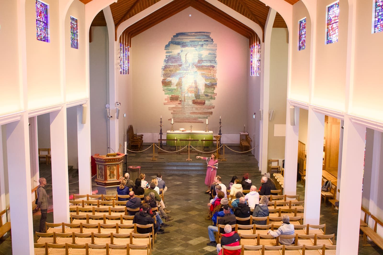 The interior of Skálholt Cathedral with stained glass windows, wooden ceiling beams, chairs, pulpit, and altar artwork.
